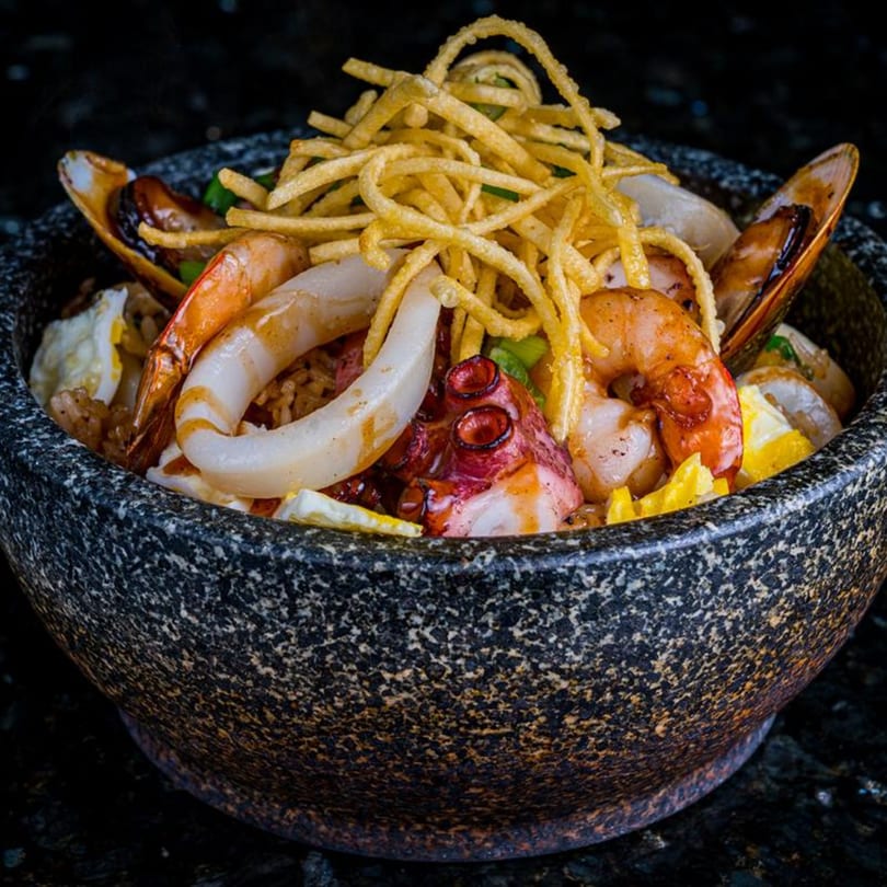 Seafood dish with shrimp, mussels, squid, and octopus, topped with crispy noodles in a stone bowl.