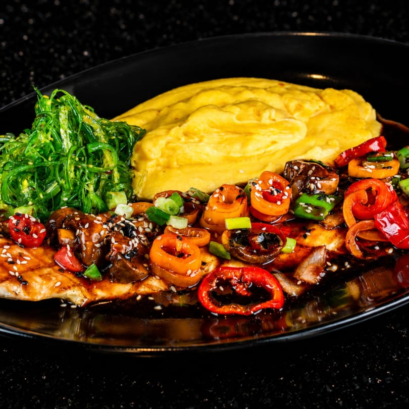 Grilled chicken with bell peppers, mushrooms, seaweed salad, and mashed potatoes on a black plate.