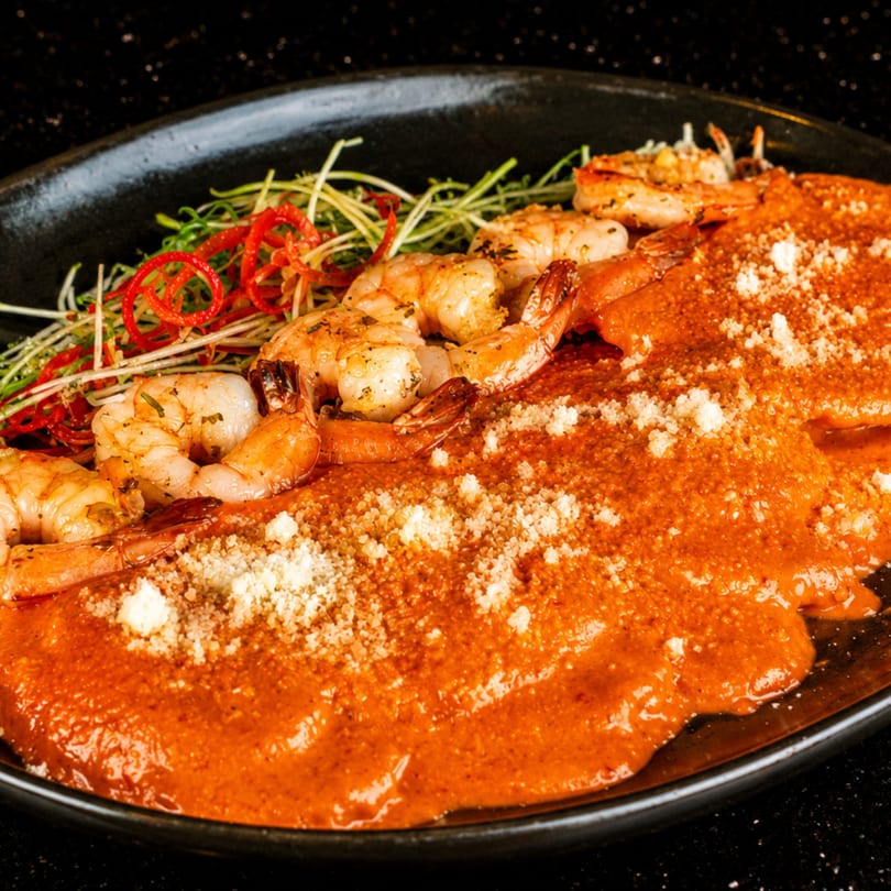 Grilled shrimp with red sauce and microgreens on a black plate.