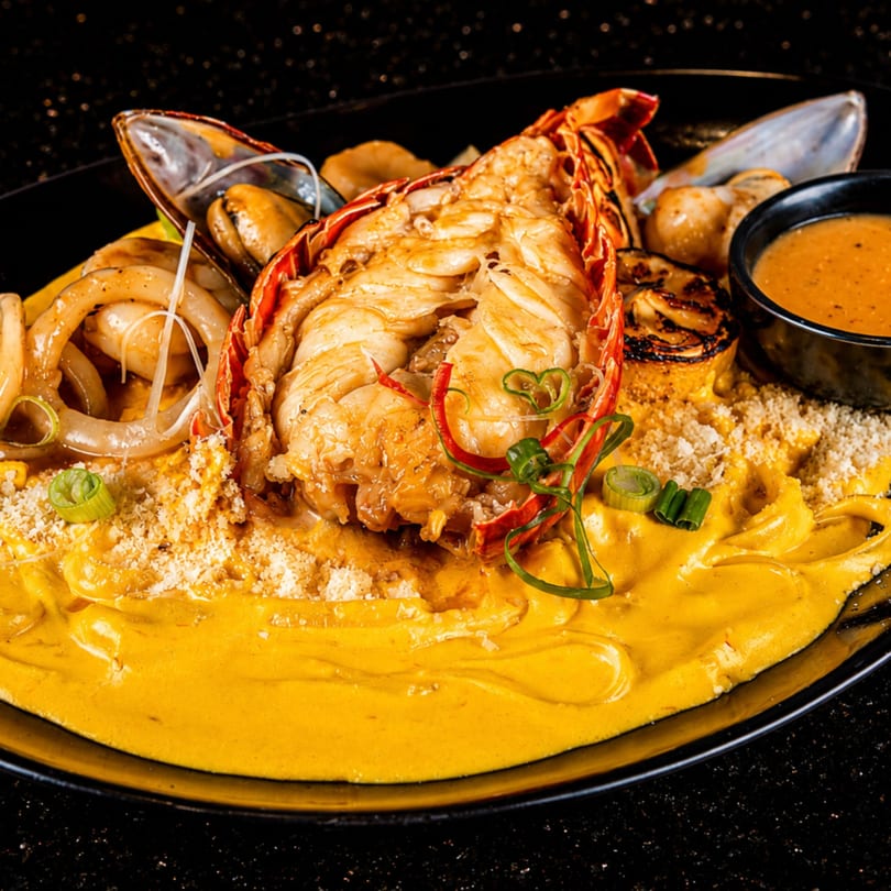 A vibrant seafood dish featuring a lobster tail, mussels, calamari, and scallops on a bed of yellow sauce.