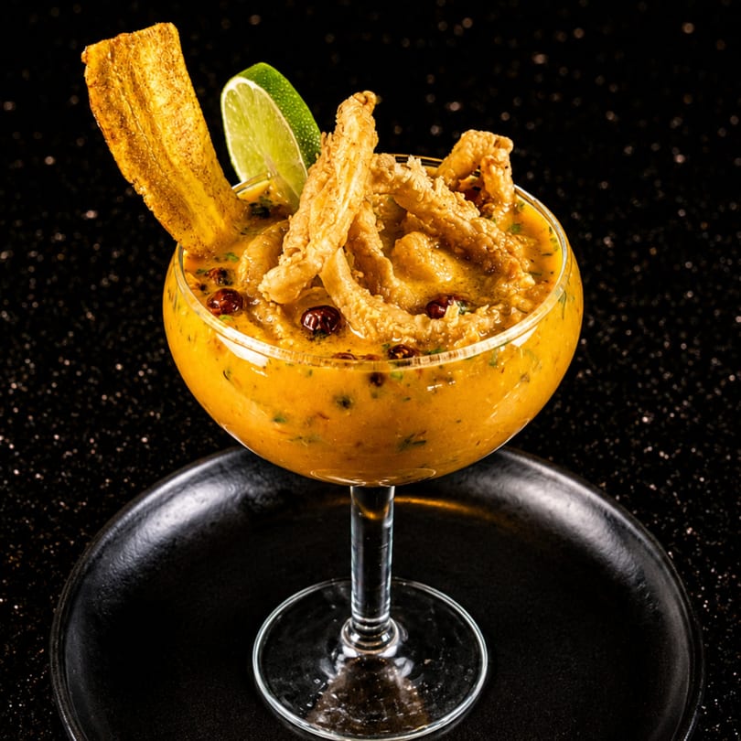 A vibrant orange ceviche in a stemmed glass, garnished with fried plantain, crispy fish, and a lime wedge.