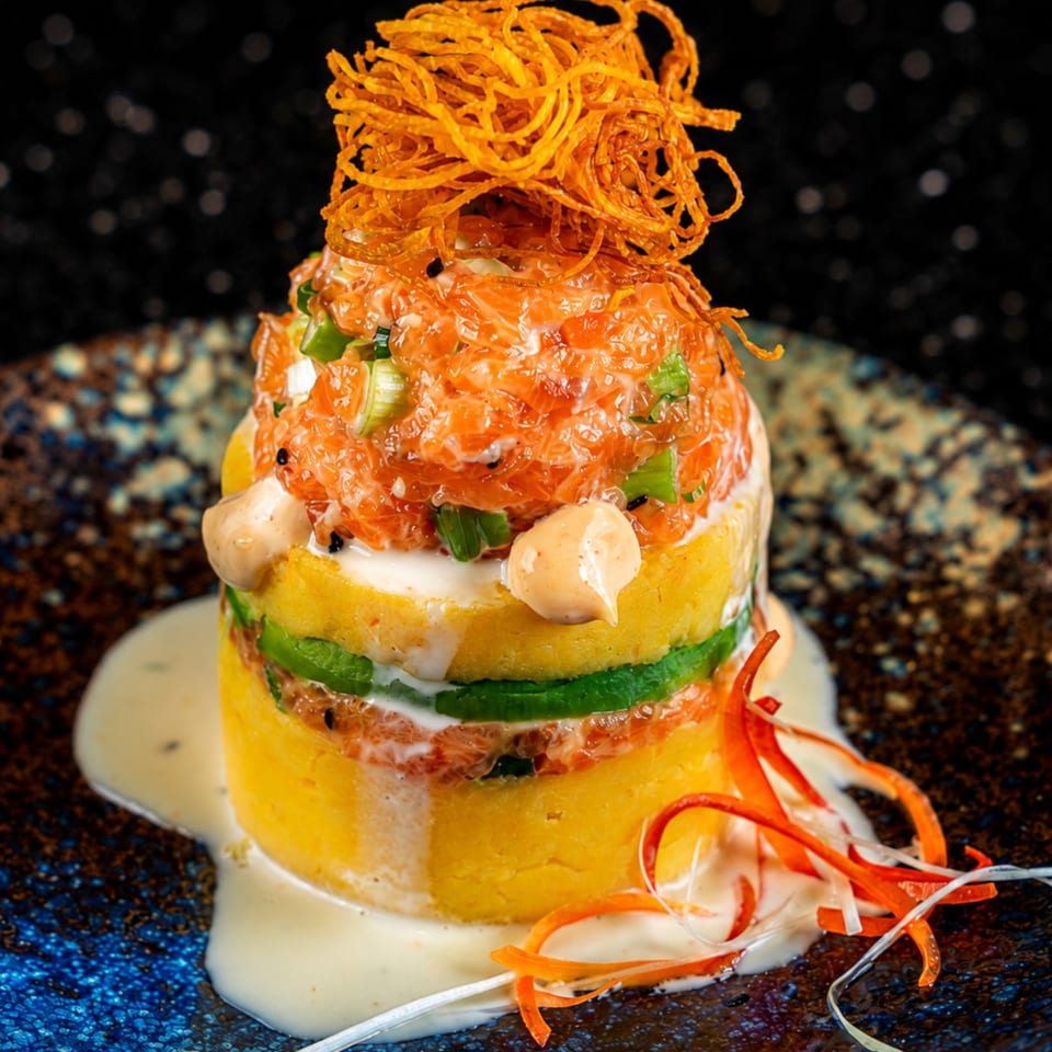 Layered salmon tartare with avocado, yellow mashed potatoes, and crispy fried onions on a dark plate.