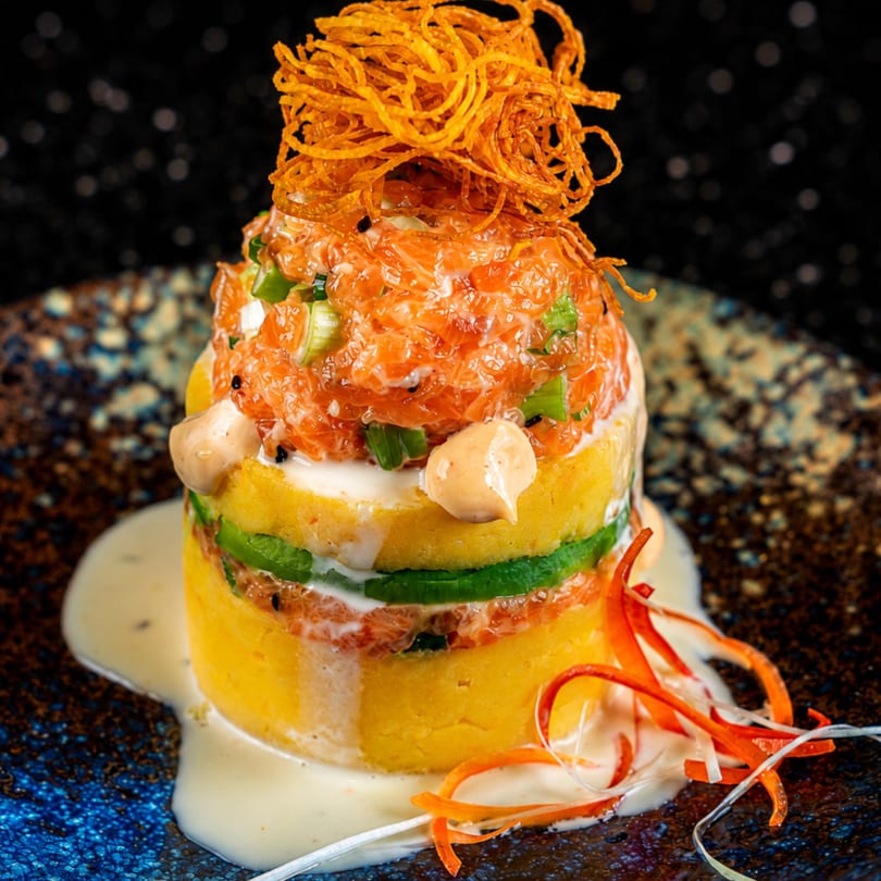 Layered salmon tartare with avocado, yellow mashed potatoes, and crispy fried onions on a dark plate.